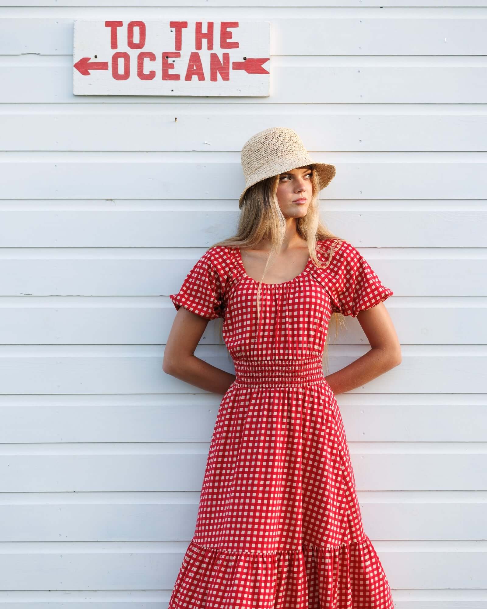 Model wearing the Sweetheart Smocked Dress by Downeast, styled with a hat, against a beach-themed backdrop.