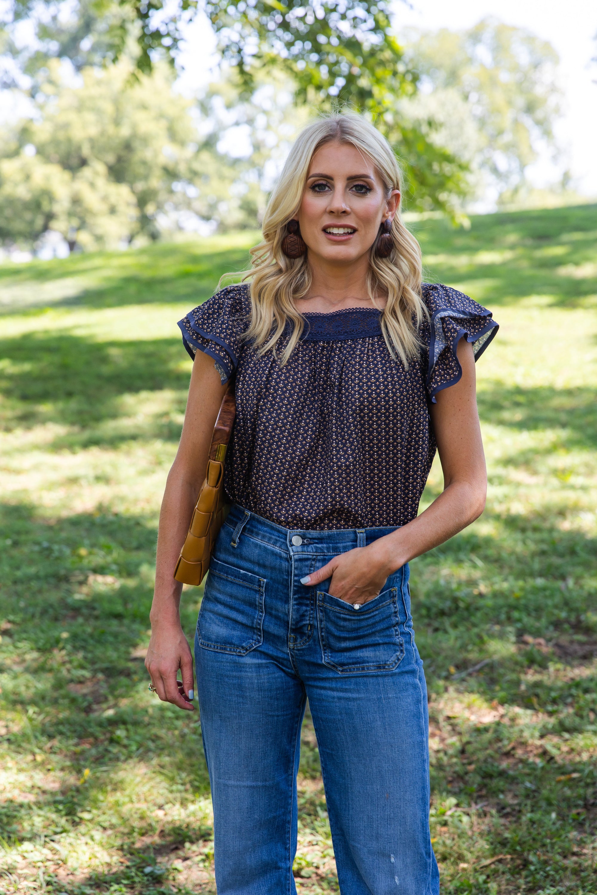 Woman wearing James Top Fall Floral Navy Lace Flutter Sleeve with high-waisted jeans in a sunny park setting.